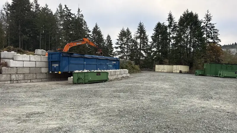 Orange excavator loading a blue bin in the Salt Spring Island transfer station drop-off yard, with green rental bins behind